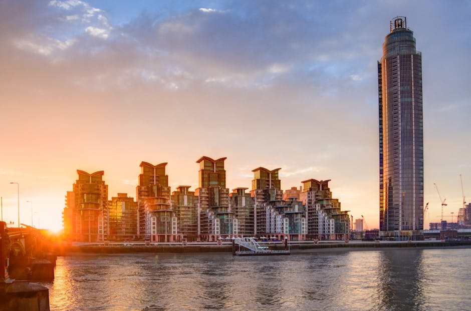 A cityscape view of Nine Elms, London, during sunset, showing a collection of modern residential buildings with a distinctive tiered, staggered design, constructed with glass and concrete materials. In the foreground, a body of water reflects the warm orange and pink hues of the sky. To the right, a tall, cylindrical skyscraper with a rounded top stands prominently above the smaller structures. The skyline includes cranes indicating ongoing construction. The scene captures the urban environment relevant to home relocation and property development, with sunlight illuminating the buildings creating soft shadows. This image is associated with the location and area serviced by Man with Van Nine Elms, focusing on removals and moving logistics within the Nine Elms district.