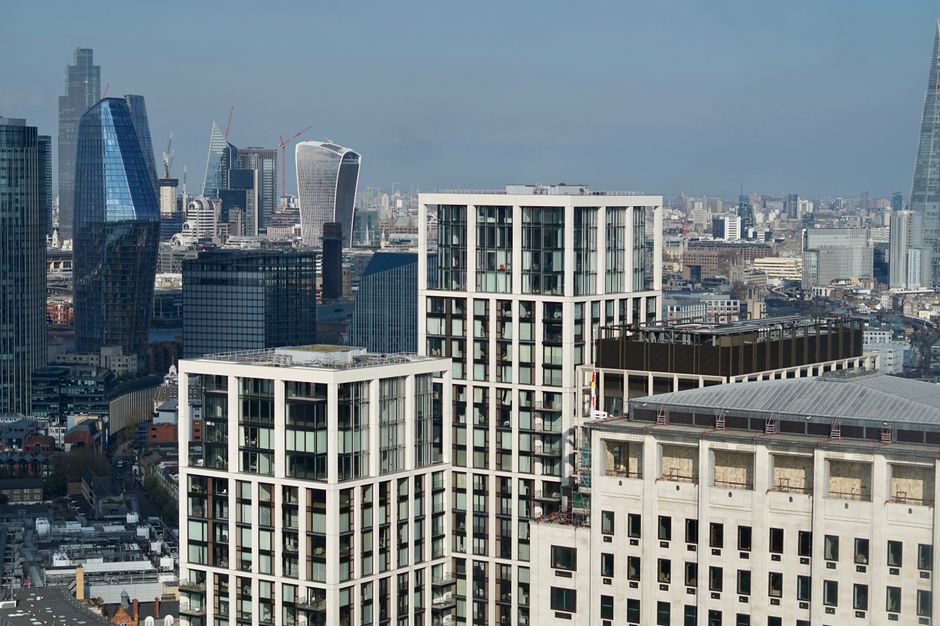 A high-angle view of a city skyline featuring modern high-rise office buildings and residential apartments near the Nine Elms area, with glass facades reflecting the sky, during daytime with clear weather. In the foreground, part of a building with a white concrete exterior and several small, rectangular windows is visible. The background shows additional skyscrapers, including some under construction with cranes on top, and a mix of older and newer structures extending into the distance. The scene captures the urban environment where home relocation or furniture transport might take place, relevant to services offered by Man with Van Nine Elms specializing in removals and moving logistics.