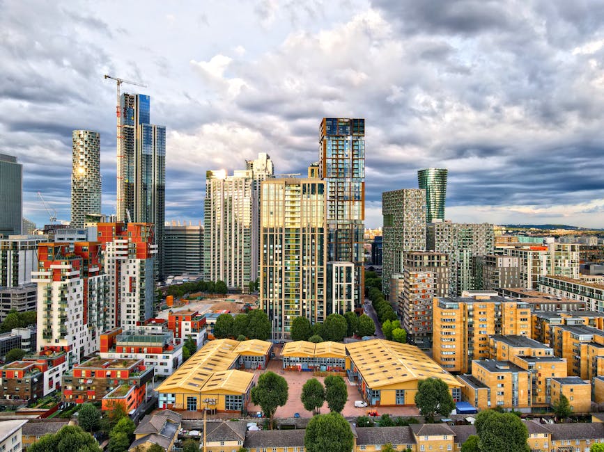 A wide view of a city skyline featuring tall modern skyscrapers with glass facades, some under construction with cranes visible, set against a partly cloudy sky. In the foreground, there are lower-rise buildings with flat roofs, surrounded by green trees and open plazas. The scene appears to be taken from an elevated vantage point, capturing a dense urban area near Nine Elms, with a mix of residential and commercial structures. The image reflects urban development and modern architecture typical of home relocation and furniture transport environments, relevant to house removals services provided by Man with Van Nine Elms.
