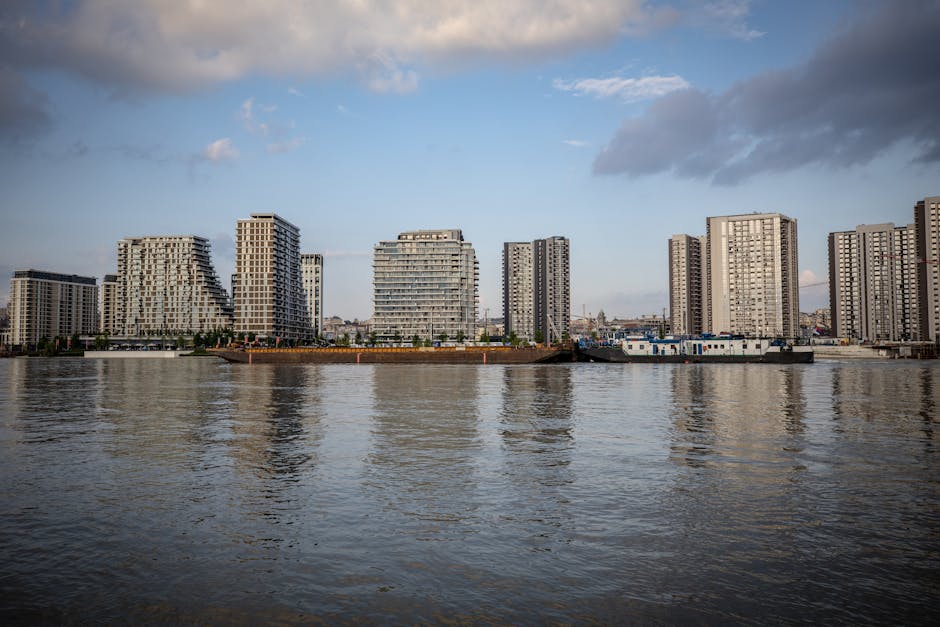 A high-angle view of a city skyline featuring modern high-rise office buildings and residential apartments near the Nine Elms area, with glass facades reflecting the sky, during daytime with clear weather. In the foreground, part of a building with a white concrete exterior and several small, rectangular windows is visible. The background shows additional skyscrapers, including some under construction with cranes on top, and a mix of older and newer structures extending into the distance. The scene captures the urban environment where home relocation or furniture transport might take place, relevant to services offered by Man with Van Nine Elms specializing in removals and moving logistics.