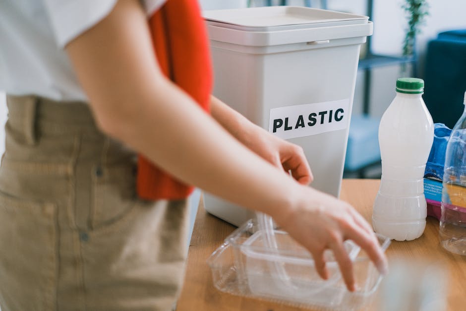 Close-up of a person placing a plastic food container into a white plastic recycling bin labeled 'PLASTIC' on a wooden table, surrounded by various bottles and packaging materials including a clear plastic bottle with a green cap and a partially visible blue plastic bag. The scene takes place indoors, with natural lighting illuminating the workspace used for packing and sorting household waste in preparation for removal or disposal. The individual is wearing light-colored pants and an orange or red top, focused on organizing items into the appropriate recycling bin as part of home relocation or moving logistics. The background features a blurred view of a living space, indicating a household environment. This image relates to the process of packing, moving, and adhering to local waste disposal rules in SW8 (Wandsworth Council), with a focus on responsible household waste separation during house removals by Man with Van Nine Elms.