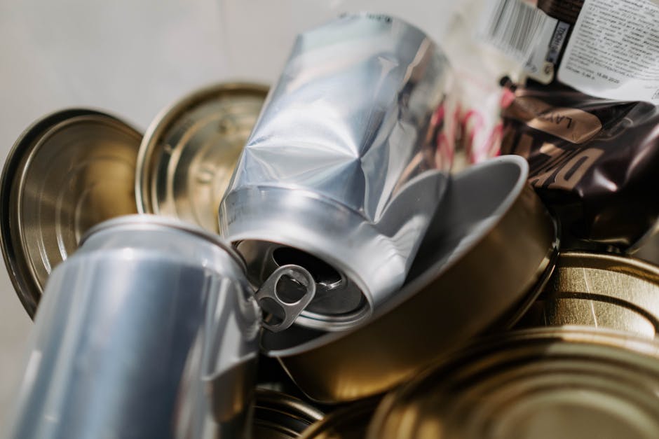 Close-up image of a collection of discarded aluminium cans, some crushed and others intact, piled together with a few plastic food containers and cardboard packaging materials. The cans are silver, with pull tabs visible, and are positioned inside a cardboard box that is partially open. In the background, additional packaging items such as food wrappers and plastic bottles can be seen, indicating household waste awaiting disposal or recycling. This scene is set in an indoor environment, possibly near an entryway or storage area, with natural or ambient lighting illuminating the waste materials. The arrangement suggests the process of consolidating household waste for collection during a home relocation or moving process, with [COMPANY_NAME] providing professional removals and logistics services, including waste disposal adhering to local disposal rules like those in SW8 (Wandsworth Council).