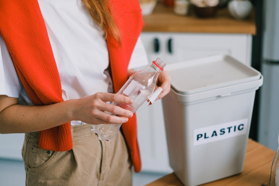 Close-up of a person placing a plastic food container into a white plastic recycling bin labeled 'PLASTIC' on a wooden table, surrounded by various bottles and packaging materials including a clear plastic bottle with a green cap and a partially visible blue plastic bag. The scene takes place indoors, with natural lighting illuminating the workspace used for packing and sorting household waste in preparation for removal or disposal. The individual is wearing light-colored pants and an orange or red top, focused on organizing items into the appropriate recycling bin as part of home relocation or moving logistics. The background features a blurred view of a living space, indicating a household environment. This image relates to the process of packing, moving, and adhering to local waste disposal rules in SW8 (Wandsworth Council), with a focus on responsible household waste separation during house removals by Man with Van Nine Elms.