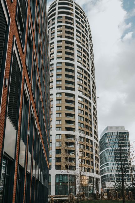 A close-up view of the exterior of a modern high-rise apartment building at Embassy Gardens, Nine Elms, featuring curved glass balconies with black metal railings arranged in a vertical line. The buil