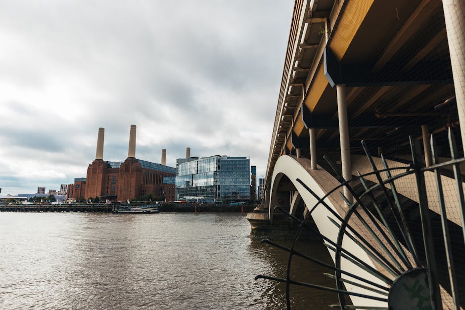 Urban scene showing the River Thames with a view of the Battersea Power Station buildings in the background, featuring three tall brick chimneys and a modern glass extension. In the foreground, part of a building structure with visible support beams, a large black fan, and a section of a tram or bus stop canopy. The pavement and street-level area are adjacent to the water, with cloudy skies overhead, suggesting overcast weather. The scene captures the area around Nine Elms, near Battersea Power Station, during daylight hours, illustrating the urban environment that Man with Van Nine Elms services for home relocation and furniture transport.
