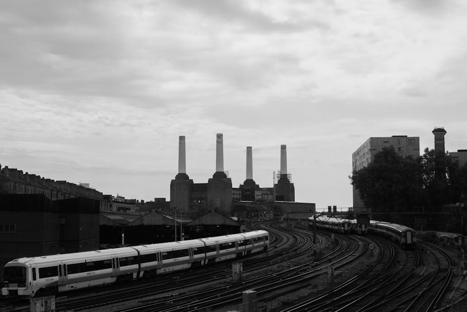 A black and white photo taken from inside or near a property near Nine Elms, showing a section of rail tracks with two modern commuter trains parked on the platforms. In the background, the iconic chimney stacks of the Battersea Power Station are visible under a cloudy sky, with some surrounding buildings and trees adding context to the urban environment. The scene captures elements relevant to home relocation and furniture transport, with the rail tracks and trains indicating logistics involved in moving belongings, and the overall setting reflecting an area undergoing redevelopment near Nine Elms, with the image associated with Man with Van Nine Elms' removals services.