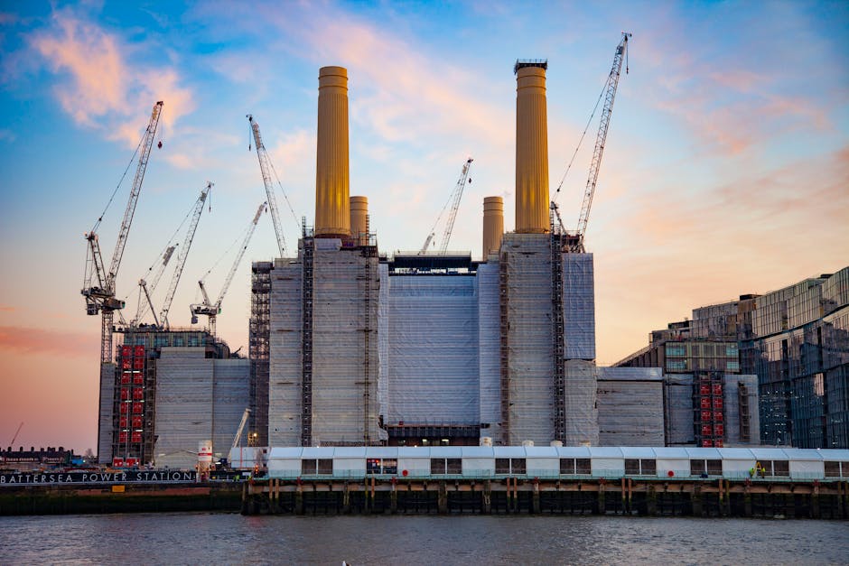 Urban scene showing the River Thames with a view of the Battersea Power Station buildings in the background, featuring three tall brick chimneys and a modern glass extension. In the foreground, part of a building structure with visible support beams, a large black fan, and a section of a tram or bus stop canopy. The pavement and street-level area are adjacent to the water, with cloudy skies overhead, suggesting overcast weather. The scene captures the area around Nine Elms, near Battersea Power Station, during daylight hours, illustrating the urban environment that Man with Van Nine Elms services for home relocation and furniture transport.