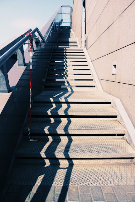 An outdoor metal staircase with non-slip treads ascending alongside a beige building, featuring black metal railings on the left side with a red and white safety chain hanging from the top. Shadows cast by the railing create a zigzag pattern on the steps, highlighting the stairwell's angle. The staircase appears to serve as an access route to upper floors or a rooftop, situated on Wandsworth Road in Nine Elms. Inside the property, partially visible at the top of the stairs, are cardboard boxes, wrapping materials, and possibly a piece of furniture, indicating a home relocation or moving process. During loading or unloading, the stairs are used for transporting packing crates and household items, aligning with services provided by Man with Van Nine Elms. The environment is well-lit by natural daylight, emphasizing the functional and practical nature of the staircase in house removals and furniture transport tasks.