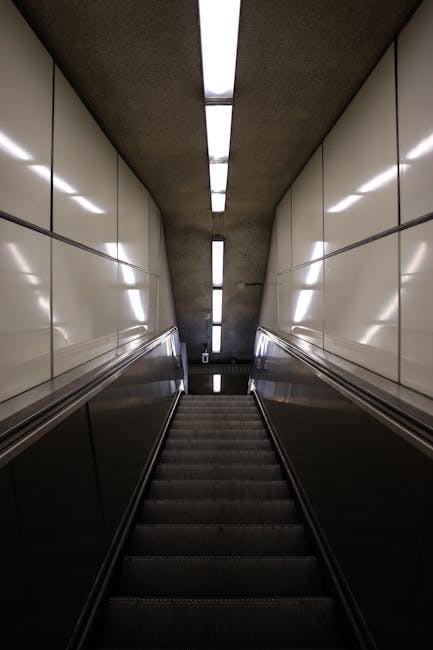 A downward view of a stairwell inside a building, showing metal and glass handrails on both sides, with beige tiled walls and a narrow strip of fluorescent lighting running along the ceiling. The stairs are made of dark material, possibly carpeted or rubberized, and lead down to an unseen lower level. The environment suggests an underground or underground-access point, with a focus on interior design typical of public transport stations or underground walkways. This image could relate to the loading or access point for a home relocation service, with [COMPANY_NAME] facilitating moves through narrow staircases or basement entries as part of their house removals and furniture transport services.