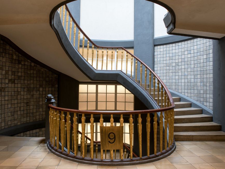 A wide view of an indoor staircase in a building with stone walls and three tall, rectangular windows at the landing allowing natural light to illuminate the space. The staircase features green-tiled
