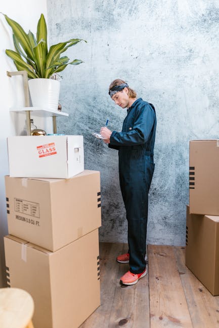 A man wearing blue coveralls and a headband stands indoors against a light textured wall, surrounded by stacked cardboard boxes of various sizes. One box has a red label that reads 'GLASS'. The man is holding a pen and clipboard, appearing to make notes during a home relocation or packing process. Several cardboard boxes are positioned on a wooden floor, with some on a low table or shelf on the left side. A large potted plant with long green leaves sits atop the shelf, adding a decorative element to the room. The scene depicts a professional packing or moving task, possibly part of a furniture transport or house removals service offered by Man with Van Nine Elms, with a focus on careful handling and organization of packing materials in preparation for a house move or office relocation.
