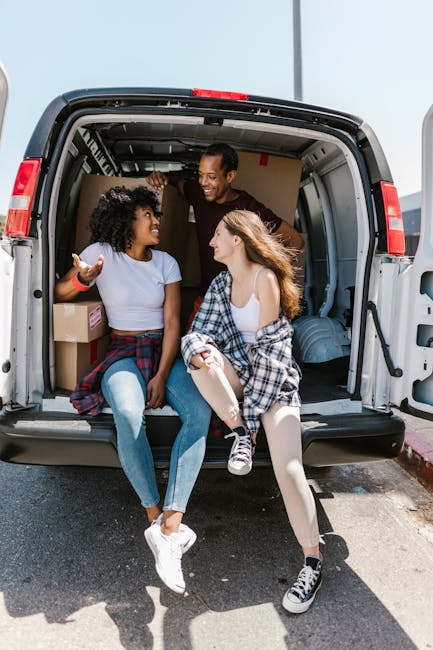 Three young adults are sitting at the open back of a grey removal van on a paved area outside a residential property. The van's interior shows stacked cardboard boxes and wrapped furniture pieces, ind