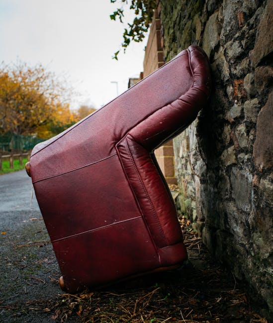A large, red leather sofa, likely a bulky piece of furniture, has fallen onto its side against a stone wall on a paved outdoor surface. The sofa's upholstery shows minor creases and wear, with the arm