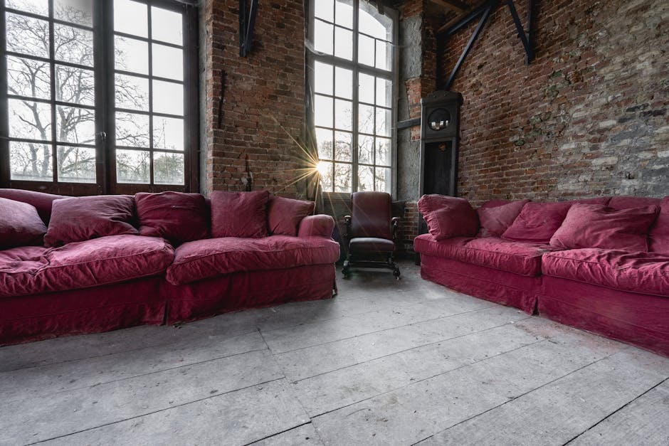 A large, red leather sofa, likely a bulky piece of furniture, has fallen onto its side against a stone wall on a paved outdoor surface. The sofa's upholstery shows minor creases and wear, with the arm