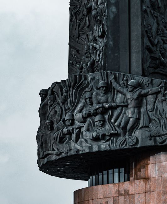 A close-up view of a large, dark metal monument featuring detailed relief sculptures of soldiers in various poses, some holding rifles and others appearing to advance or support, mounted on a cylindrical base with a reddish-brown tiled surface. The background shows an overcast sky, with the monument situated outdoors. The detailed carvings depict scenes of military personnel, reflecting themes of remembrance or historical significance, and the lighting emphasizes the texture and depth of the metalwork, which may be part of a memorial or commemorative installation, relevant to house removals and moving services by Man with Van Nine Elms during related relocation projects.