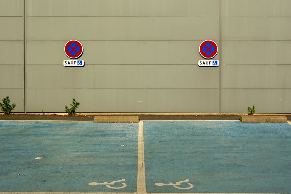 A view of a parking area featuring two designated disabled parking spaces marked with white wheelchair symbols on the blue asphalt surface. Behind the parking spaces, there is a beige wall with two prominent circular no parking signs, each with a red border, a blue background, and a red cross, indicating no parking except for disabled individuals, as shown by the small blue wheelchair icons and the word 'SAUF' underneath. Small green plants are growing between the parking spaces and the wall. The scene is evenly lit, with no vehicles or people present, providing a clear view of the parking restriction signs and layout, relevant for home relocation logistics and vehicle access management for [COMPANY_NAME]’s removals services.