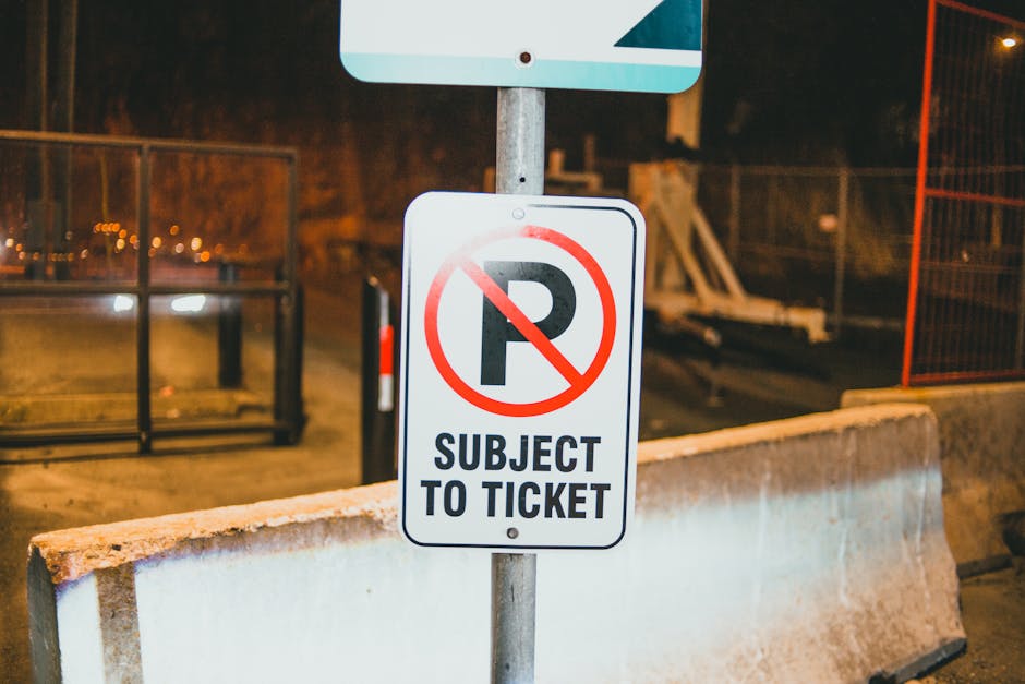 A close-up image of a rectangular parking sign mounted on a metal pole, situated behind a concrete barrier on a pavement at night. The sign features a bold black capital letter 'P' crossed out with a red circle and diagonal line, accompanied by the text 'SUBJECT TO TICKET' beneath in black capital letters. The setting includes orange safety fencing and construction materials in the background, illuminated by streetlights, indicating an active work zone. The sign is positioned at the entrance to an area where parking is restricted, relevant to moving and housing relocation logistics managed by Man with Van Nine Elms during home removals or furniture transport activities.